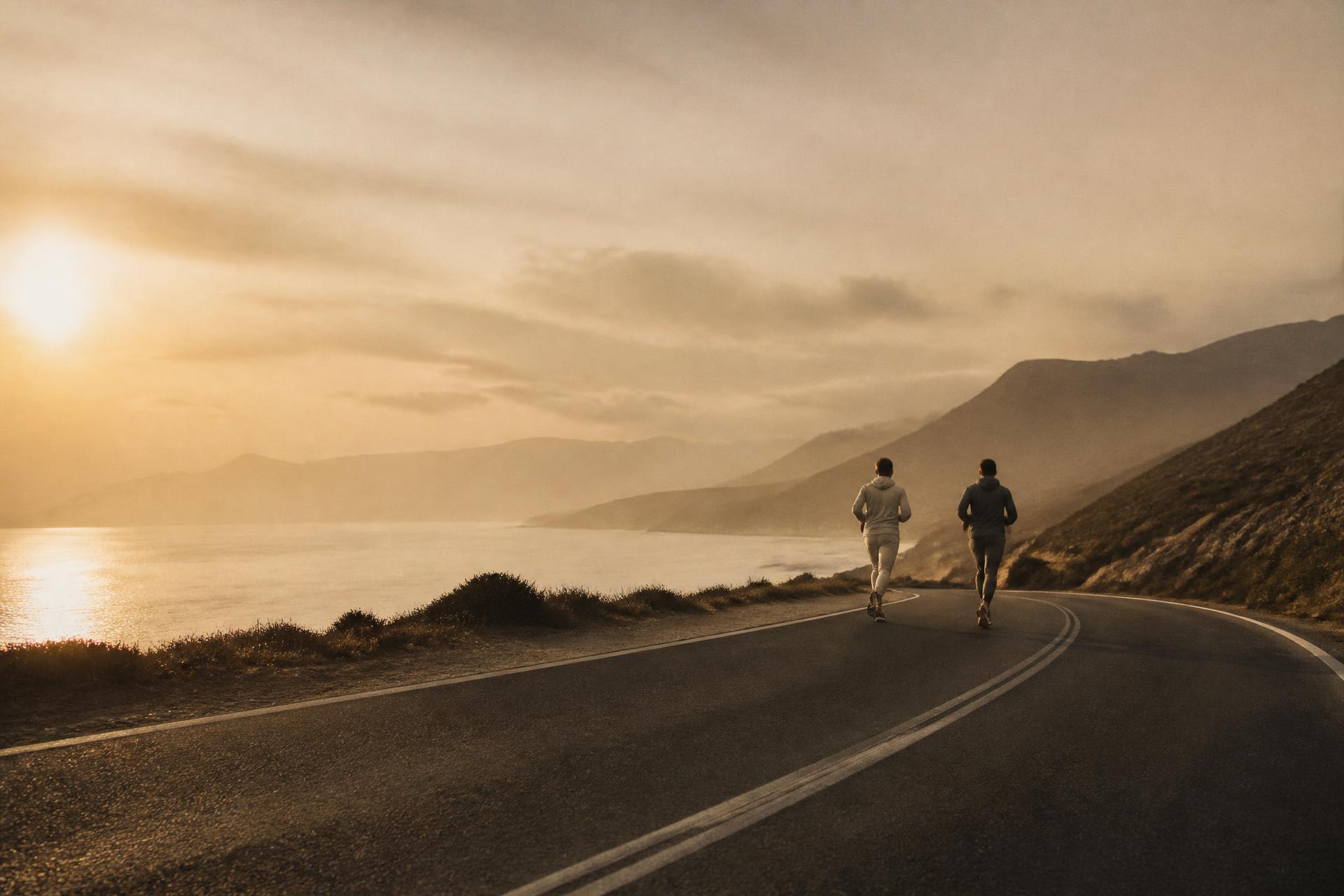 Duas pessoas correndo numa estrada litorânea vazia ao pôr do sol, com o mar e montanhas ao fundo
